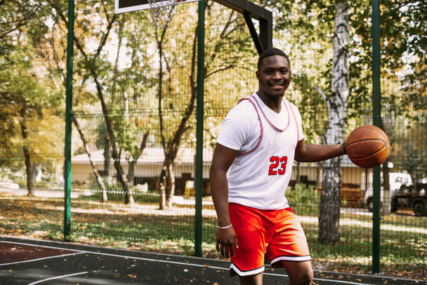 Retrato de un apuesto joven negro sosteniendo una pelota de baloncesto en una cancha de baloncesto. Tómese un descanso durante su entrenamiento. Retrato deportivo de moda - Foto, Imagen