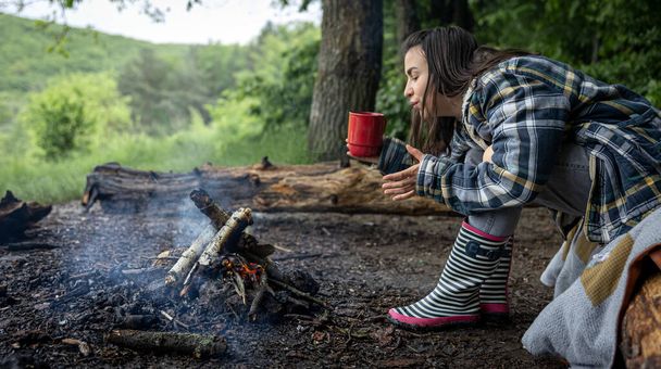 An attractive girl with a cup in her hand warms up near a fire in the forest. - Foto, Bild