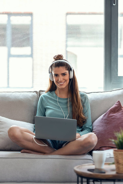 Shot of confident young woman using her laptop while listening to music with headphones sitting on a couch at home. - Фото, изображение