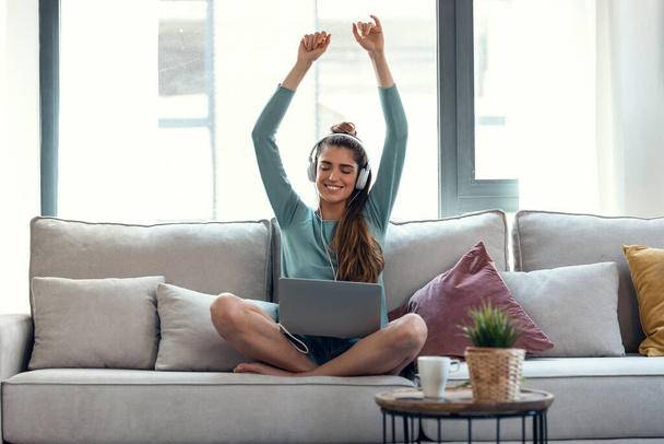 Shot of motivated young woman dancing and listening to music with her laptop while sitting on sofa at home. - Photo, Image