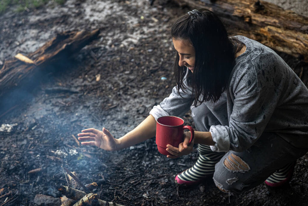Egy lány csészével a kezében tűzet gyújt, hogy melegen tartsa.. - Fotó, kép