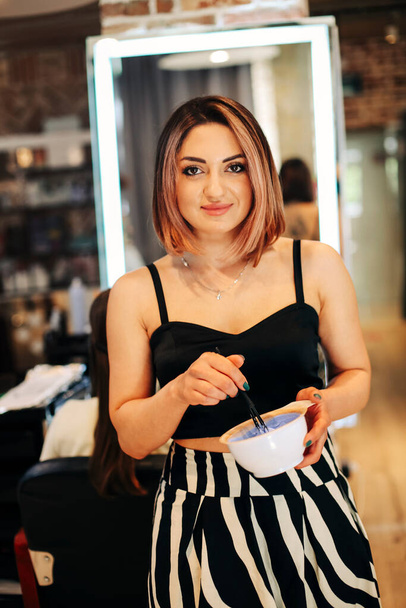Young hairdresser mixing hair dye in bowl while standing near mirror and woman in modern beauty salon - Photo, Image