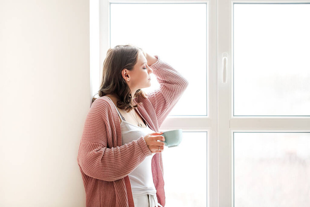 Young female with cup of hot drink touching head and relaxing near window in sunny morning at home - 写真・画像