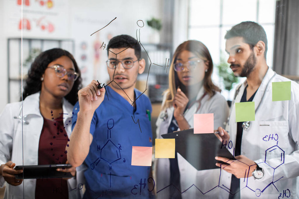 Team of doctors, discussing in front of glass wall, writing formulas, using notes and stickers - Fotoğraf, Görsel