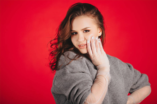 Junge Frau in stylischen durchscheinenden Handschuhen berührt Gesicht und blickt vor rotem Hintergrund in die Kamera - Foto, Bild