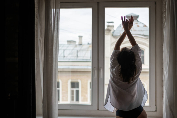 Beautiful sensual young woman with long hair in a white shirt standing at the window back - Φωτογραφία, εικόνα