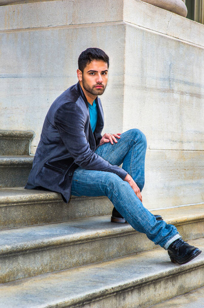 Habillé d'un blazer en laine violet foncé, d'un jean bleu et de chaussures en cuir noir, un jeune beau garçon avec un peu de pain et de moustache est assis sur les marches d'un immeuble de bureaux, vous regardant attentivement - Photo, image