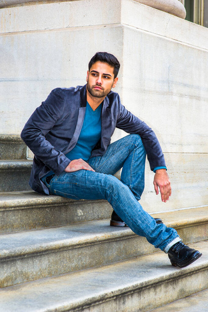 In einem dunkelvioletten Wollblazer, blauen Jeans und schwarzen Lederschuhen sitzt ein junger gutaussehender Mann aus dem Nahen Osten mit Brot und Schnurrbart auf den Stufen vor einem Büro und schaut sich nachdenklich um. - Foto, Bild
