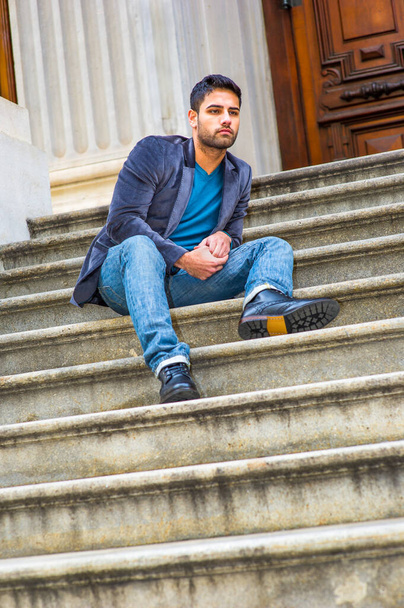 Habillé d'un blazer en laine violet foncé, d'un jean bleu et de chaussures en cuir noir, un jeune homme du Moyen-Orient avec du pain et une moustache est assis sur les marches, regardant autour et souriant - Photo, image