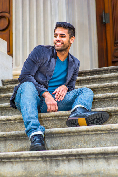 Habillé d'un blazer en laine violet foncé, d'un jean bleu et de chaussures en cuir noir, un jeune homme du Moyen-Orient avec du pain et une moustache est assis sur les marches, regardant autour et souriant. - Photo, image