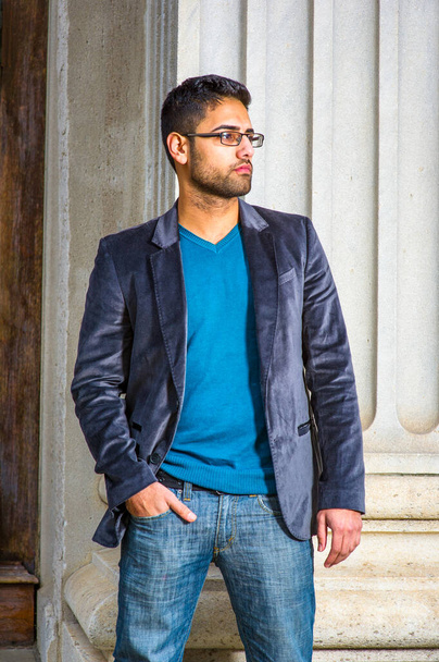 Vestido em um blazer de lã roxo escuro, suéter e jeans, usando óculos, um jovem estudante universitário com pão e bigode está parado perto de uma coluna e esperando por você - Foto, Imagem