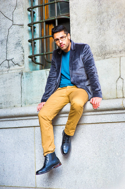 Dressing in a dark purple woolen blazer, blue sweater, dark yellow pants and leather boot shoes, wearing glasses, a young guy with bread and mustache is sitting by an old fashion style window, looking at you - Foto, Imagen
