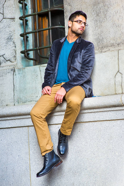 Dressing in a dark purple woolen blazer, blue sweater, dark yellow pants and leather boot shoes, wearing glasses, a young guy with bread and mustache is sitting by an old fashion style window, looking away - Photo, Image