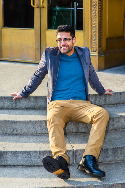 Dressing in a dark purple woolen blazer,  dark yellow pants and leather boot shoes, wearing glasses, a young professor with bread and mustache is sitting on steps, smiling - Photo, Image