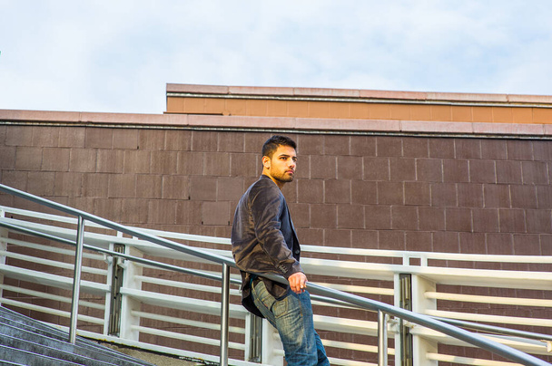 Dressing in dark purple woolen blazer and jeans, a young guy with beard and mustache is standing against a metal railing on stairs, under sunshine of sunset, thinking - Foto, imagen