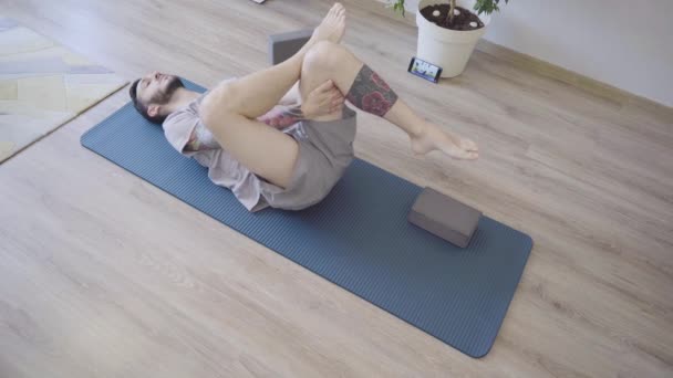 Young man practicing Pavanamuktasana yoga indoors at home - Footage, Video