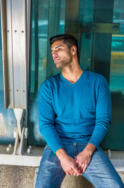 Dressing in a blue sweater and jeans , a young handsome guy with beard and mustache is sitting on the corner of a structure against a glass wall, sad - Foto, Imagem