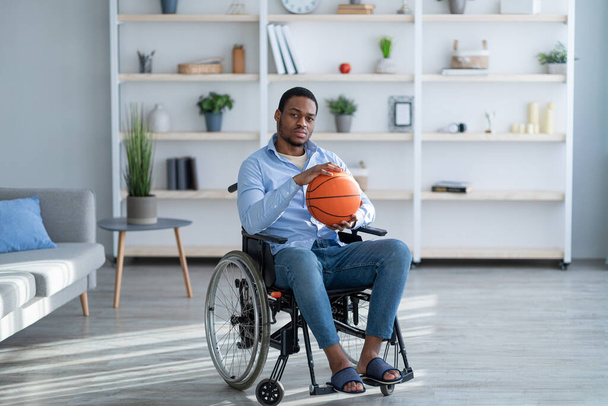 Disabled former basketball player in wheelchair holding ball with sadness, cannot play game, feeling depressed at home - Фото, изображение