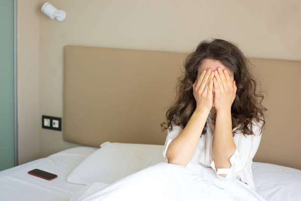 Zestresowana młoda kobieta ściskająca głowę dłońmi, cierpiąca na ból głowy, siedząca w łóżku w białej piżamie po ciężkim śnie rano. Uczucie choroby.Napięcie lub migrena problemy zdrowotne.Kopiuj przestrzeń - Zdjęcie, obraz