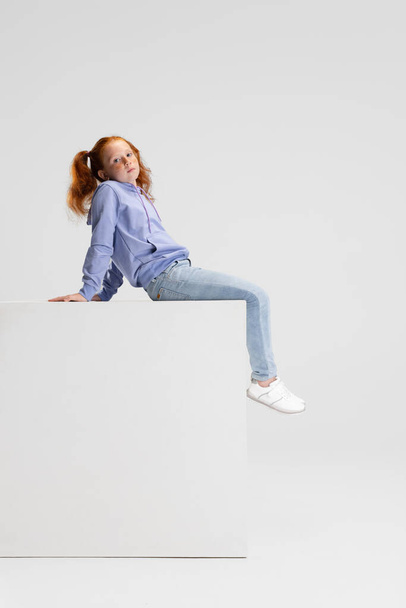 One cute red-headed girl in casual clothes sitting on big box isolated on white studio background. Happy childhood concept. Sunny child. Looks happy, delighted - Foto, imagen