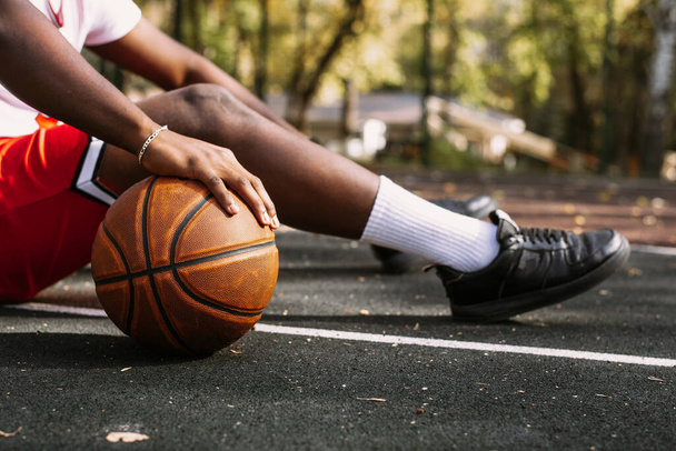 Крупный план черного молодого человека, держащего баскетбольный мяч и сидящего на баскетбольной площадке. Отдохните во время тренировки - Фото, изображение