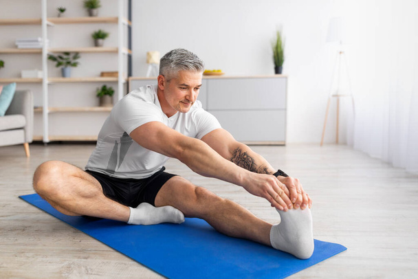 Entrenamiento en casa. Deportivo hombre maduro estirando su pierna en la estera de yoga en la sala de estar, espacio libre - Foto, Imagen