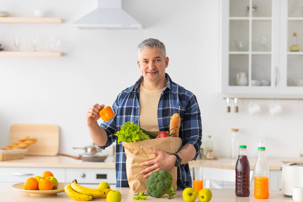 Happy mature man enjoying organic healthy food, sorting vegetables with eco package in kitchen interior, copy space - Foto, Bild