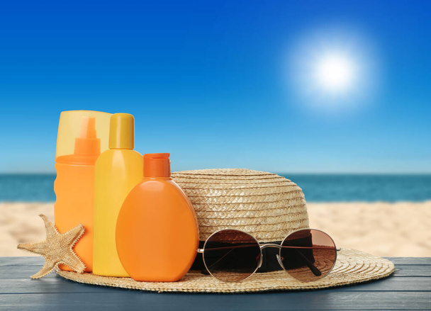 Diferentes botellas de productos de protección solar de piel y accesorios de playa en mesa de madera azul contra el paisaje marino. Espacio para el diseño - Foto, imagen