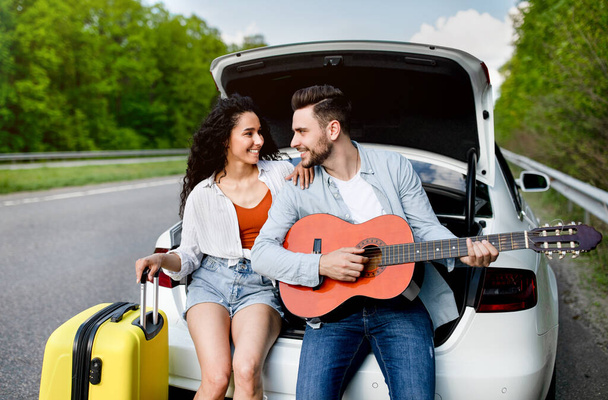 Millennial άνθρωπος και η φίλη του που ταξιδεύουν με αυτοκίνητο, κάθεται στο πορτ-μπαγκάζ του αυτοκινήτου με βαλίτσα και κιθάρα, δημιουργώντας μουσική - Φωτογραφία, εικόνα