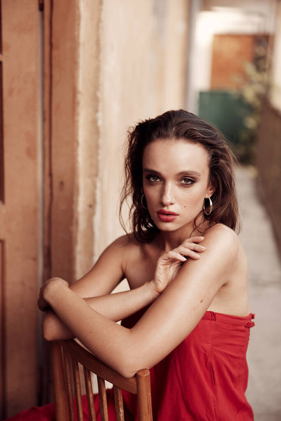 Charismatic brunette in a long red dress with beautiful facial features. sitting on a chair on the balcony in a small Spanish street. Painted red lips and incredibly beautiful brown eyes - Foto, Bild