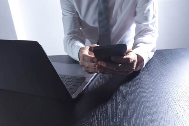 Close-up of the hands of a well-dressed man in a white shirt looking at his smartphone, using a laptop on a desk. - Photo, image
