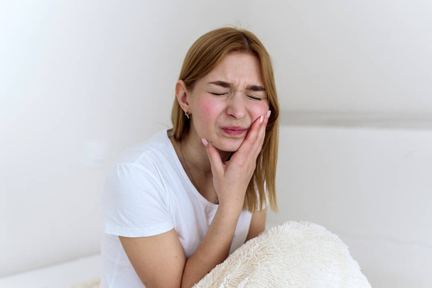 Κοντινό πλάνο μιας νεαρής γυναίκας που υποφέρει από πονόδοντο ενώ κάθεται στο κρεβάτι. Γκρο πλαν φωτογραφία ενός άνδρα που αγγίζει το μάγουλό της και στρίβει τα χείλη της λόγω πόνου στα δόντια - Φωτογραφία, εικόνα