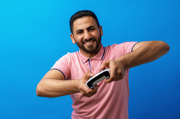 Junger arabischer Mann im rosa T-Shirt spielt Videospiele vor blauem Hintergrund - Foto, Bild