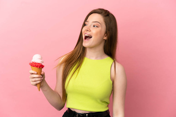 Ragazza adolescente con un gelato alla cornetta su sfondo rosa isolato ridendo in posizione laterale - Foto, immagini