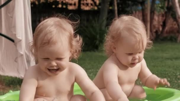 Pequeños gemelos divertidos bebé rubio jugando agua en la piscina de goma inflable de plástico infantil - Metraje, vídeo