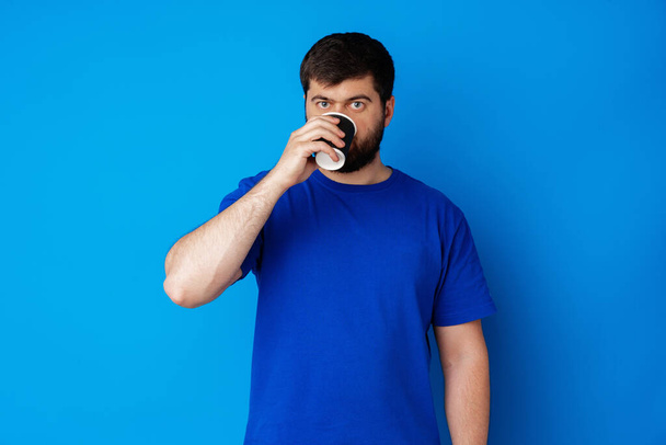 Portrait of a young bearded man drinking coffee against blue background - Foto, Bild
