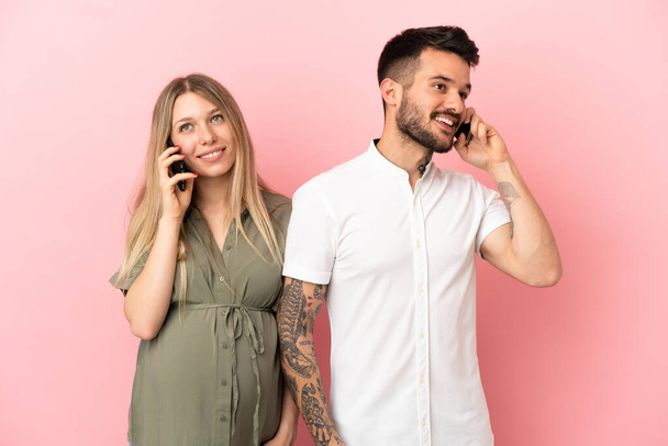Schwangere Frau und Mann vor isoliertem rosa Hintergrund im Gespräch mit Handy - Foto, Bild