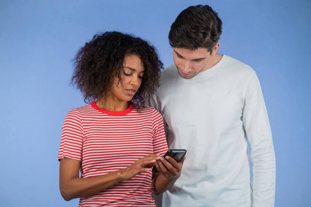 Young interracial couple using mobile device. African woman talking about apps and explains to man how to use application. Blue studio background - Foto, afbeelding