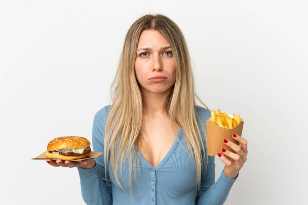 Young blonde woman holding fried chips and cheeseburger over isolated background - Photo, Image