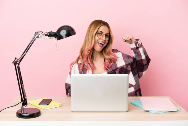 Jovem estudante mulher em um local de trabalho com um laptop sobre fundo rosa celebrando uma vitória - Foto, Imagem