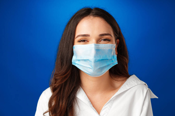 Young beautiful brunette woman wearing medical face mask against blue background - Фото, изображение