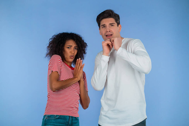 Frightened interracial couple afraid of something and looks into camera with big eyes full of horror on blue wall background. African american woman and white man. - Photo, Image