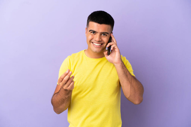 Jovem homem bonito usando telefone celular sobre fundo roxo isolado fazendo gesto de dinheiro - Foto, Imagem