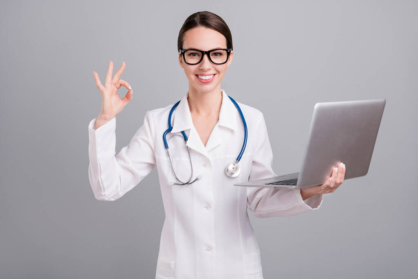 Photo of charming sweet doc woman dressed white coat spectacles typing modern device showing okey isolated grey color background - Foto, Imagen