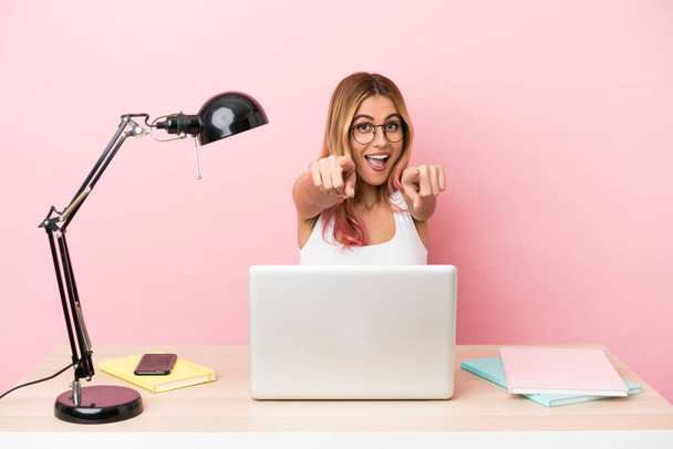 Młoda studentka w miejscu pracy z laptopem na różowym tle zaskoczona i wskazująca z przodu - Zdjęcie, obraz
