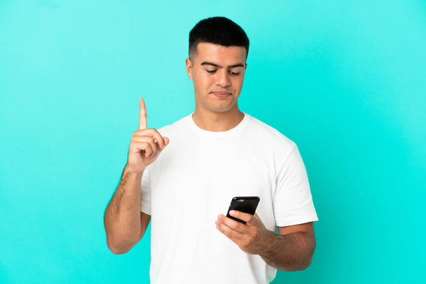 Joven hombre guapo sobre fondo azul aislado usando el teléfono móvil y levantando el dedo - Foto, Imagen