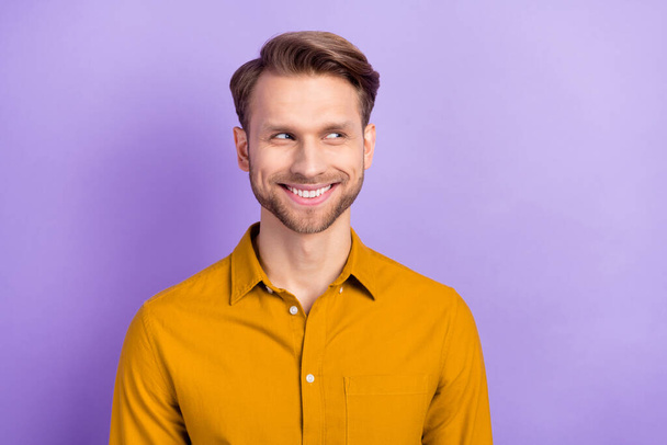 Фото смешного хитрого молодого джентльмена, одетого в желтую рубашку, улыбающегося, глядя на пустое пространство изолированного фиолетового цвета фона - Фото, изображение
