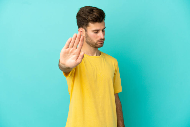 Young caucasian handsome man isolated on blue background making stop gesture and disappointed - Photo, Image