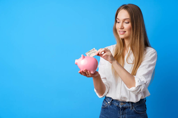 Beautiful smiling young woman with piggy bank on blue background - Fotoğraf, Görsel