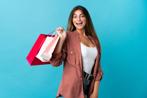 Mavi arka planda izole edilmiş genç beyaz bir kadın alışveriş torbaları tutuyor ve gülümsüyor. - Fotoğraf, Görsel